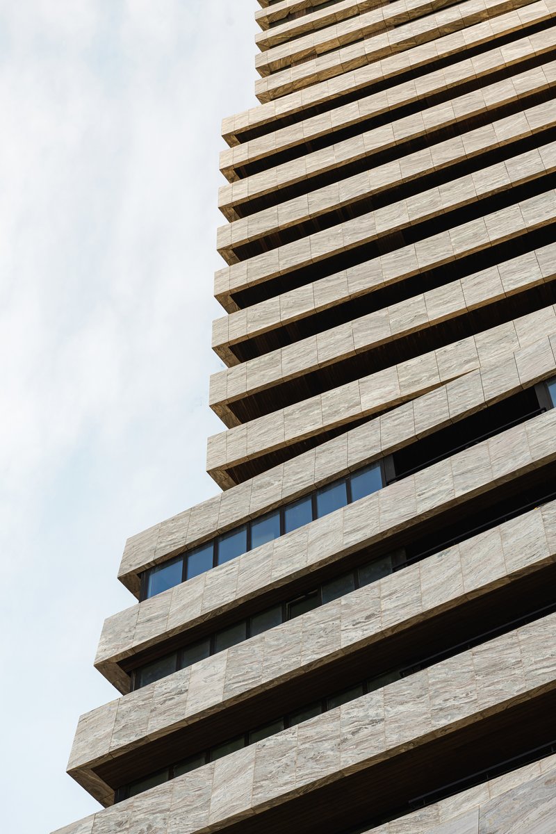 Upward view of stacked balconies with deep soffits and horizontal stone cladding against cloudy sky
