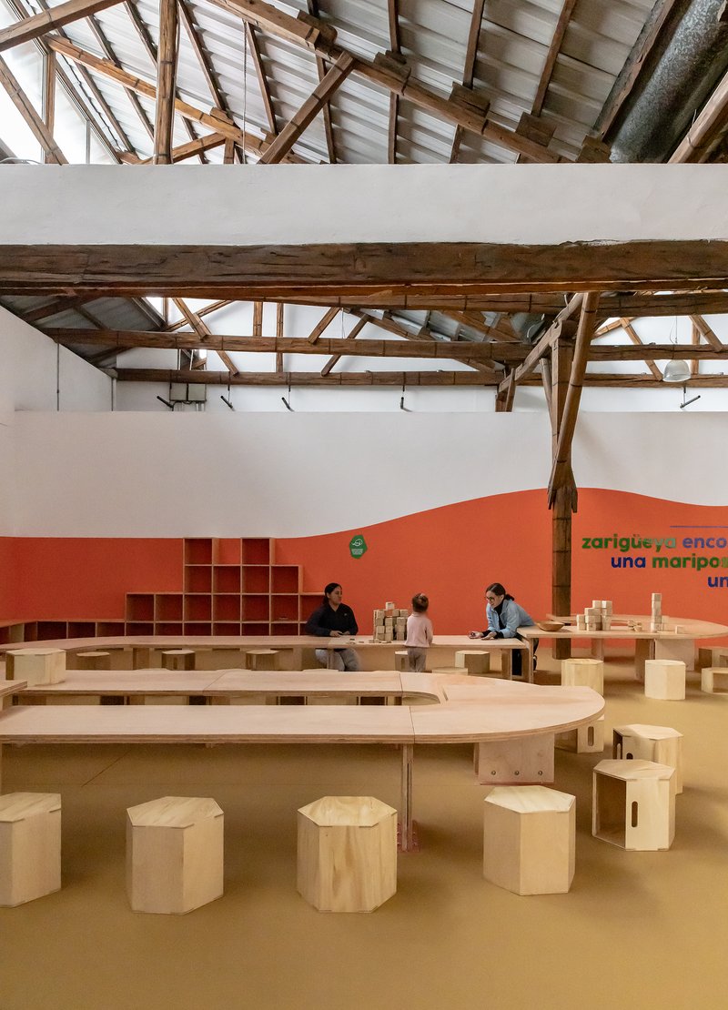 Exhibition hall with curved plywood benches and coral-painted wall beneath exposed timber trusses