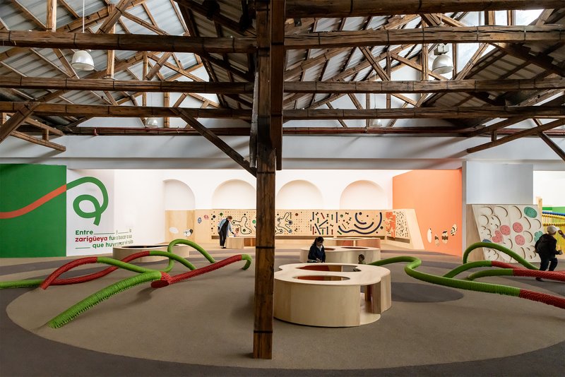 Central timber column with curved plywood seating and visitors in the open exhibition hall