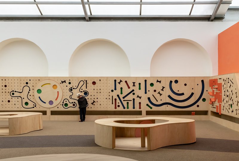 Plywood pegboard wall with circular cutouts and interactive elements facing curved timber seating benches
