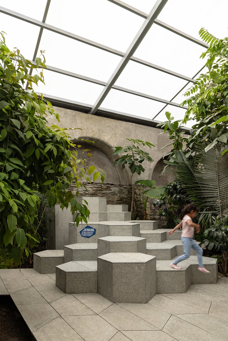 Hexagonal concrete block staircase beneath a glazed skylight surrounded by green plants and a child climbing