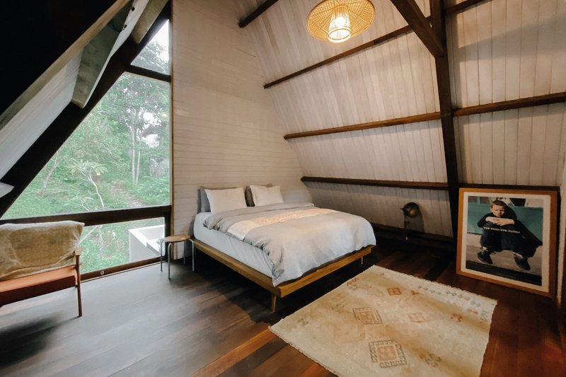A-frame bedroom with exposed dark timber rafters and triangular glazing overlooking dense green forest