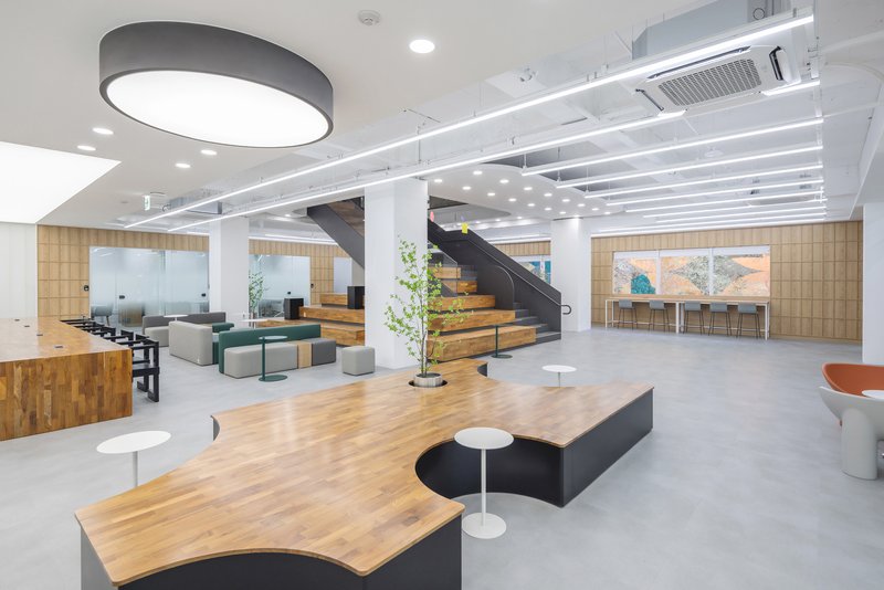 Open office interior with timber benches, black steel staircase and white columns under linear ceiling lights