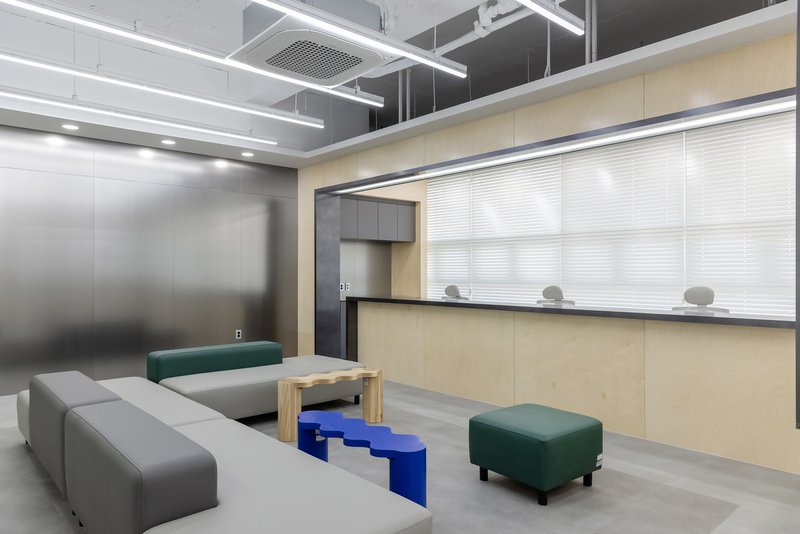 Waiting area with light wood service counter, translucent roller blinds and modular grey and green ottomans