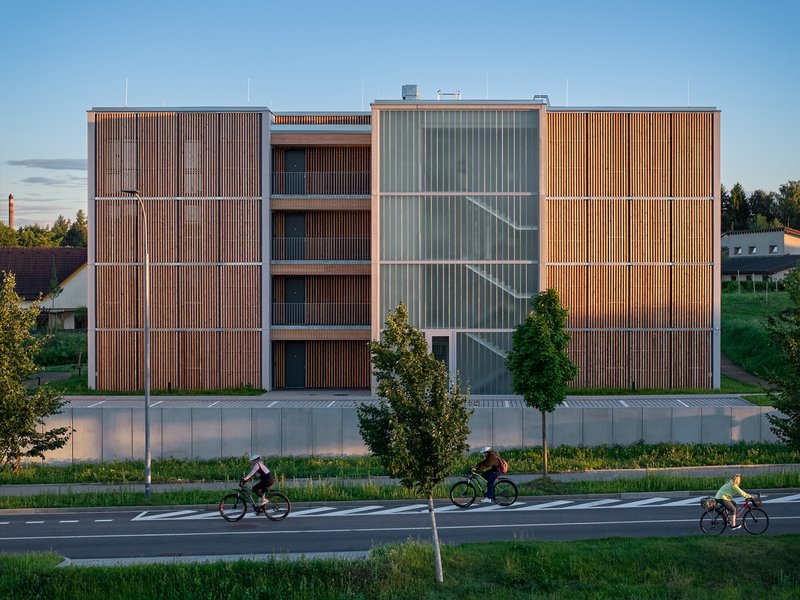Street facade with alternating timber and translucent panel screens as three cyclists pass at dusk