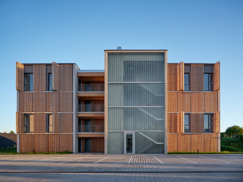 Street elevation showing timber slat volumes flanking a central translucent corrugated stair tower at dusk