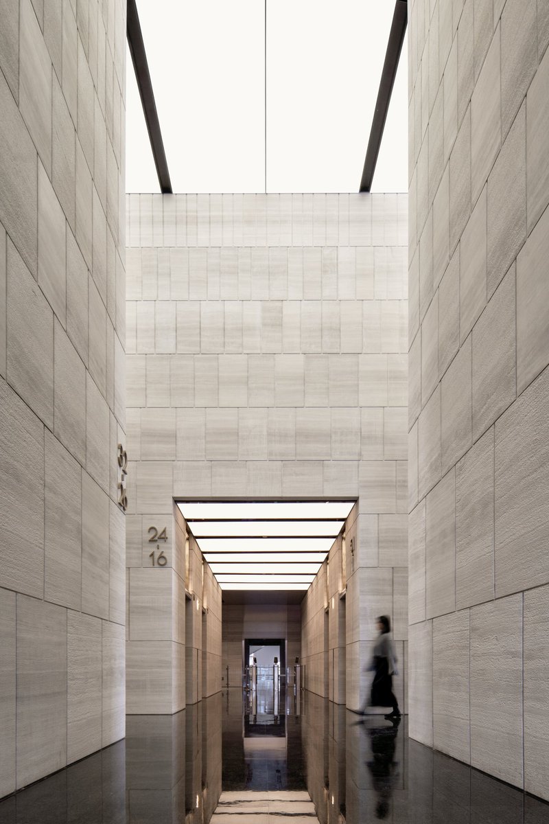 Tall atrium void lined with stone panels, a figure passing through the corridor below