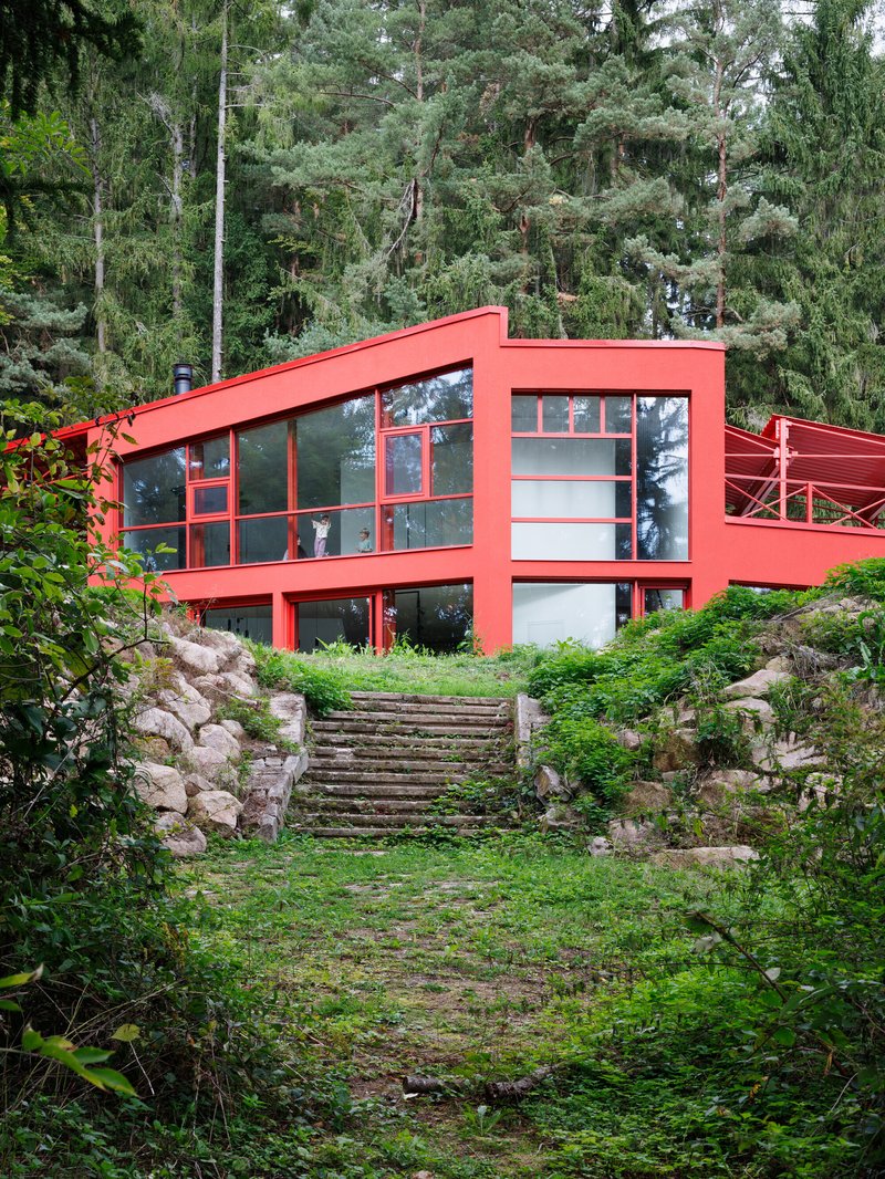 Red steel-framed facade with floor-to-ceiling glazing above a moss-covered stone stairway