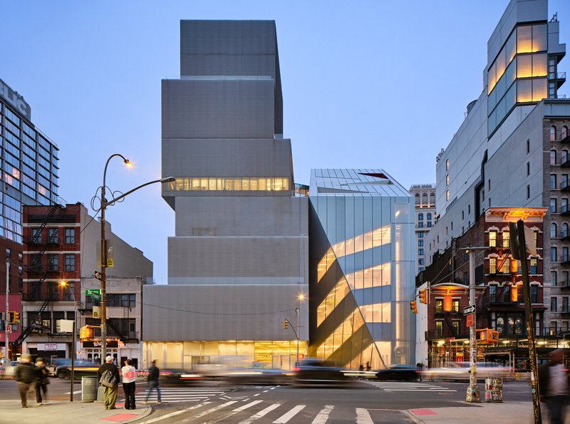 Street view of the illuminated metal facade with angled glass volumes at dusk