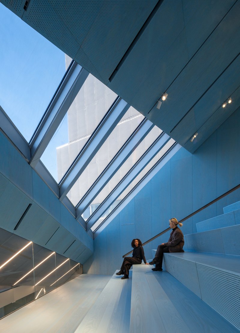 Interior stairwell with perforated metal panels, angled skylight, and two visitors seated on steps