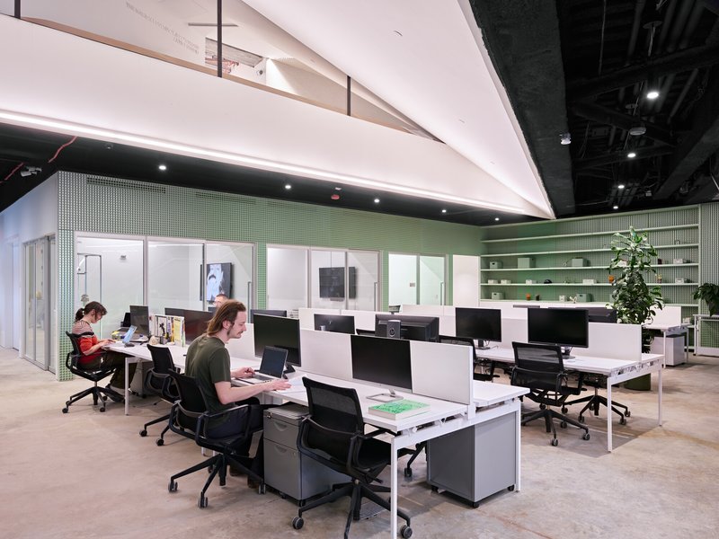 Open-plan office space with white workstations, green tiled walls and shelving under exposed industrial ceiling