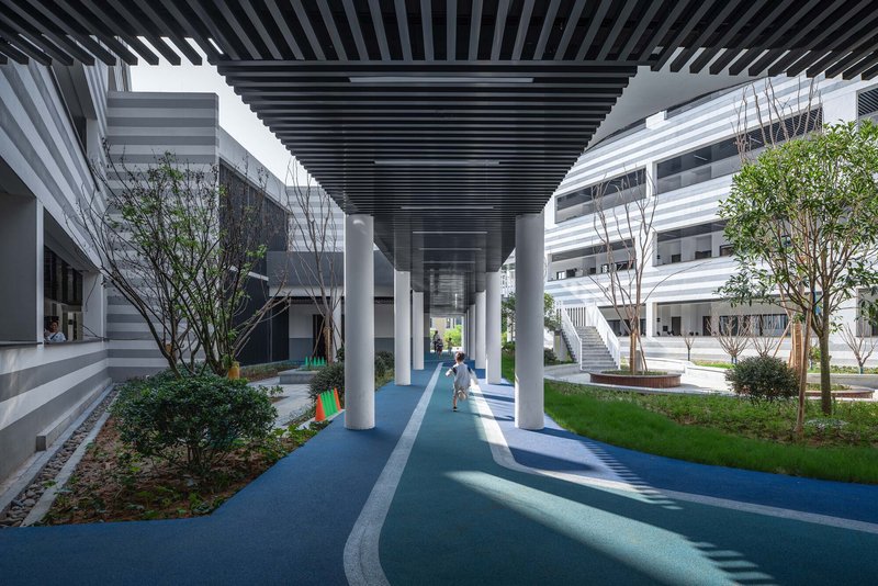 Covered walkway with slatted ceiling casting shadows on blue and grey paving beside planted trees
