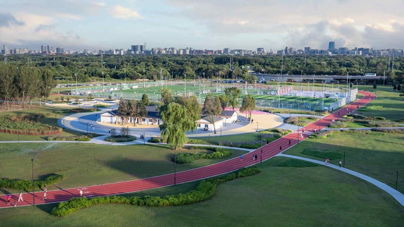 Winding red track alongside curved white path through parkland with distant city skyline under cloudy sky