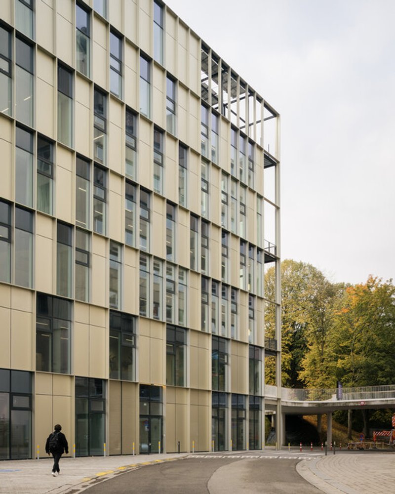 Side facade: yellow-tinted glazing panels in a gridded envelope, covered walkway, figure crossing, autumn trees, overcast Belgian sky