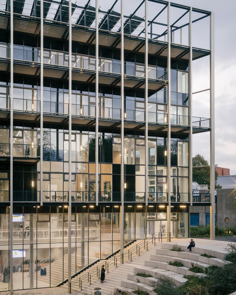 Glazed facade at dusk: full-height black steel frame wrapping the building, the sunken entry amphitheatre stepping down, figure seated at the edge, warm interior light