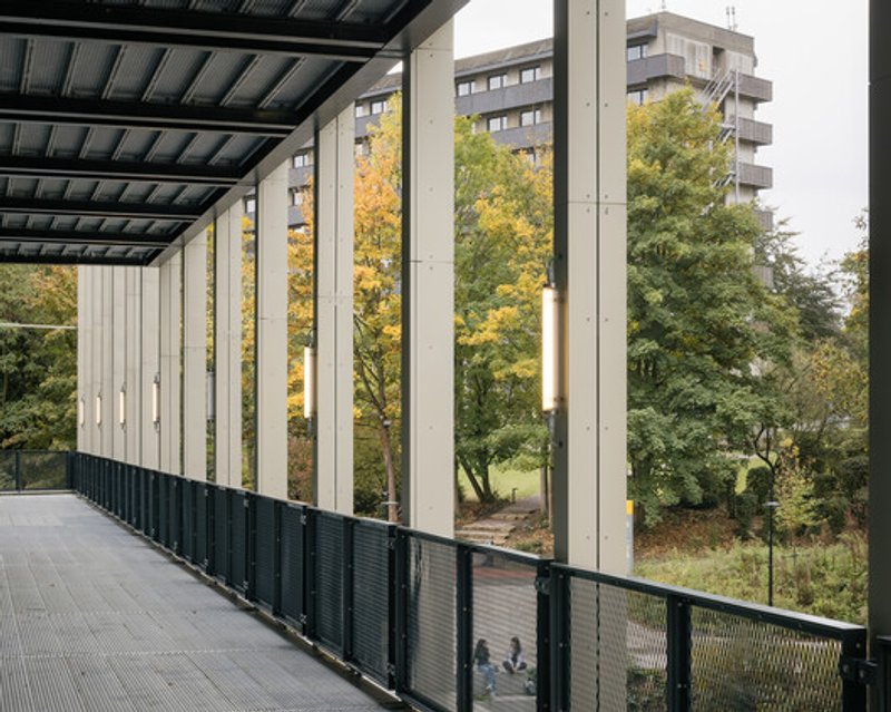 Circulation gallery: glass walkway on a mezzanine edge overlooking the atrium, slender white columns, painted black steel soffit, trees visible outside