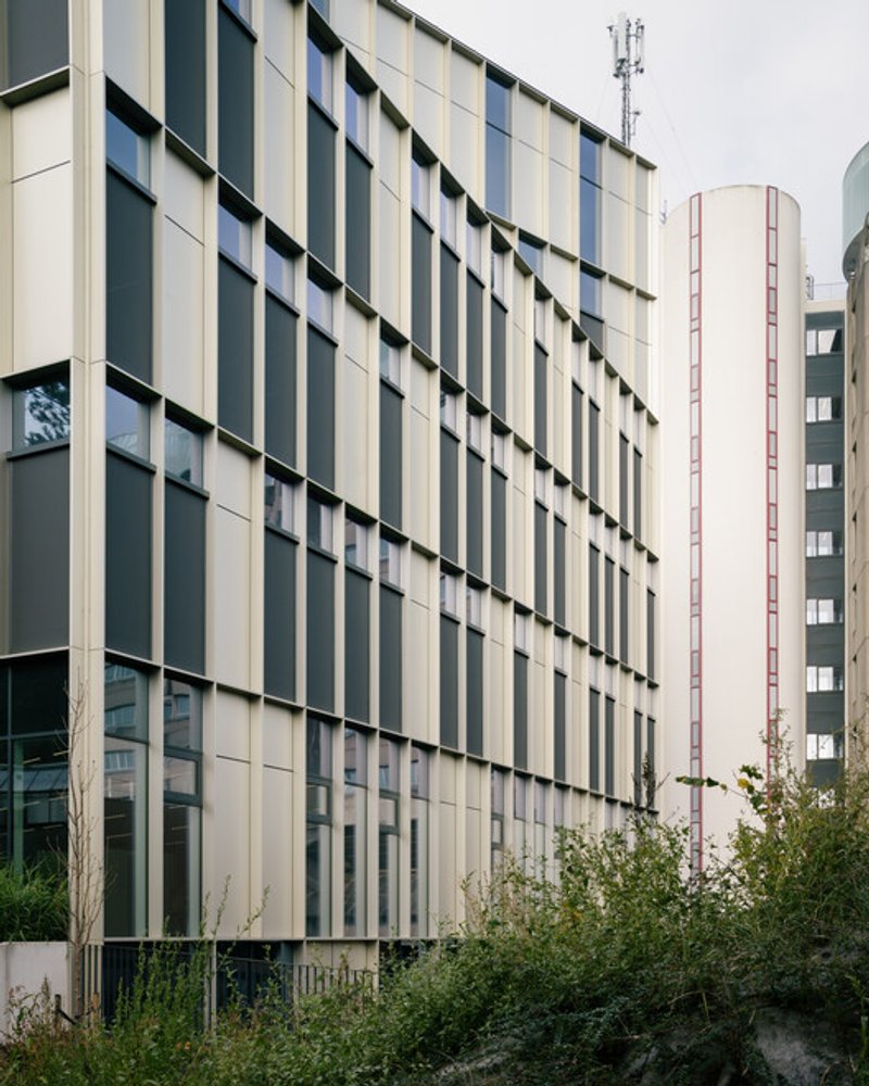 Alternate facade view: yellow-tinted glazing and black panels in an irregular grid, neighbouring tower in the background, planted buffer at the base