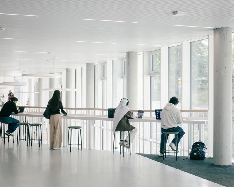 Upper reading area: bright white concrete reading room with a continuous desk along the glazed facade, stools, students working under a mushroom ceiling column, trees outside