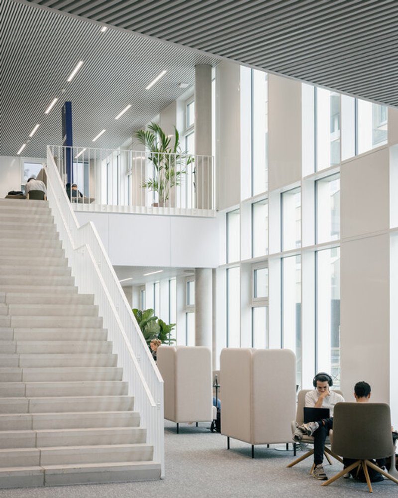 Main atrium stair and lounge: two runs of a white stair meeting a lounge with upholstered privacy booths, double-height glazing, planted trees in the volume