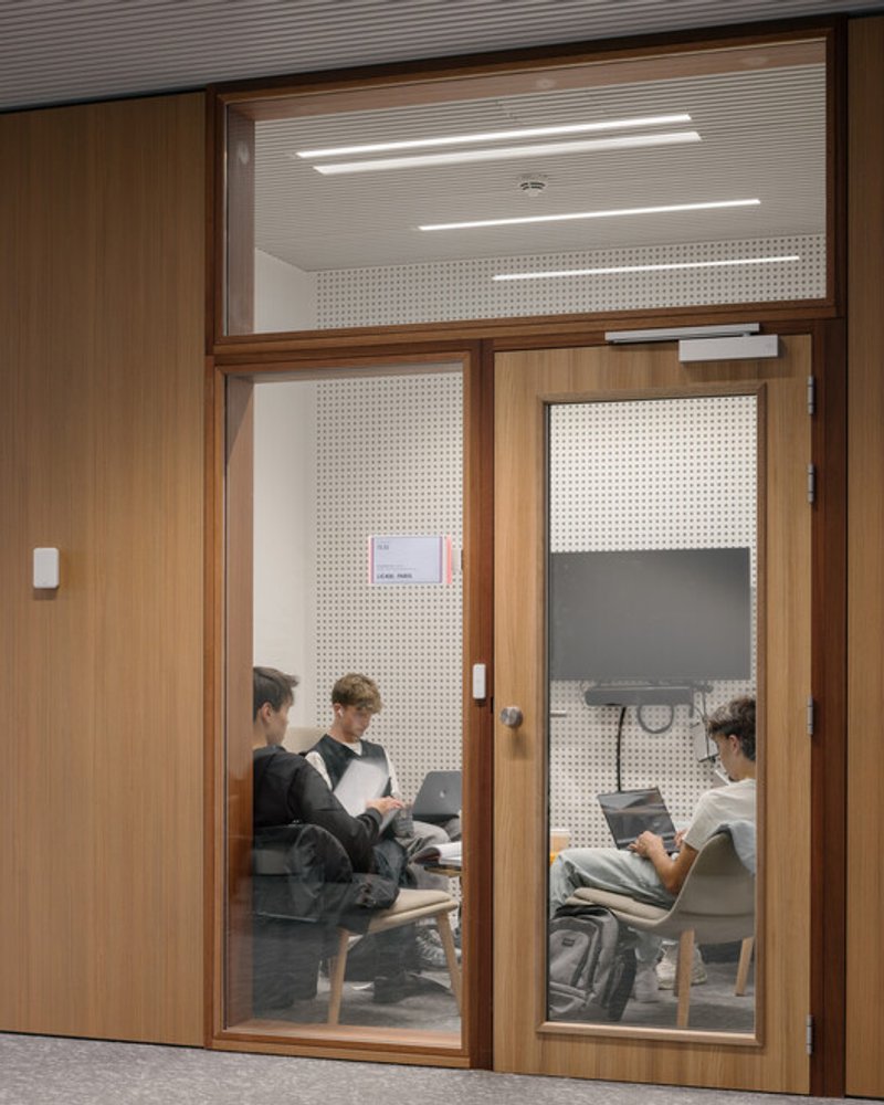 Group study room: timber door and frame, perforated acoustic panel, two students working inside at a timber table with laptops