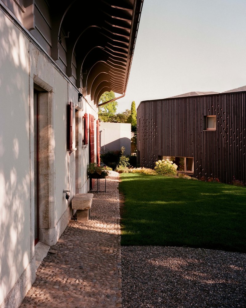 Courtyard pathway with cobblestone paving between a white masonry wall and a timber-clad volume in afternoon light