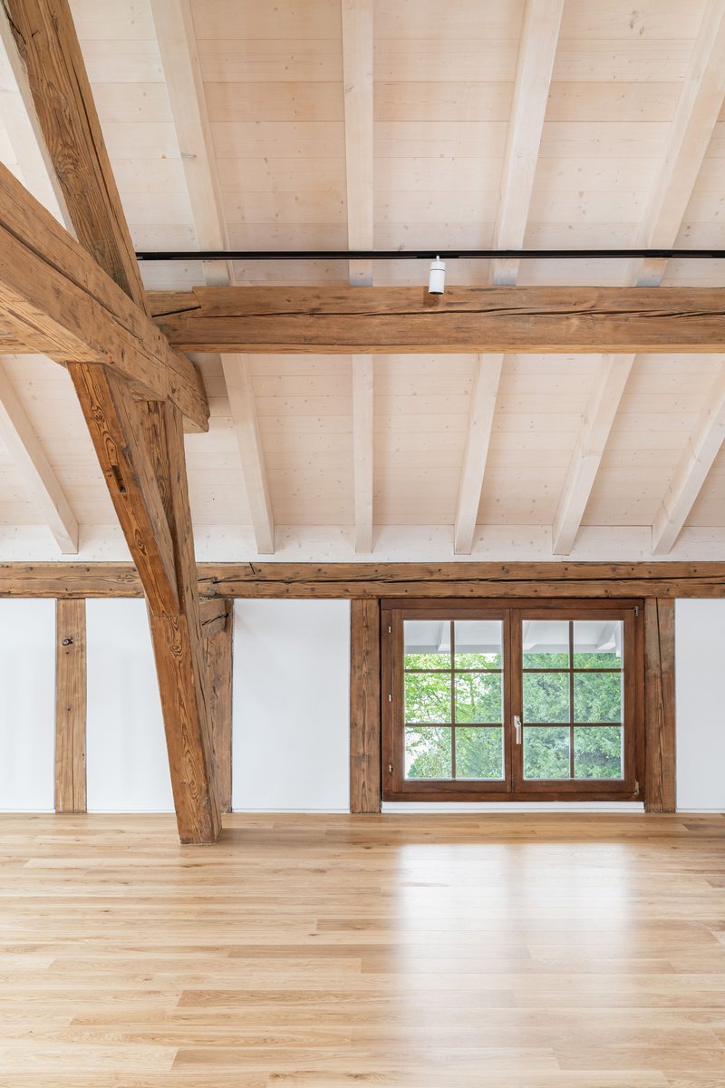 Upper floor room with exposed timber frame and wood plank ceiling overlooking green trees through gridded window