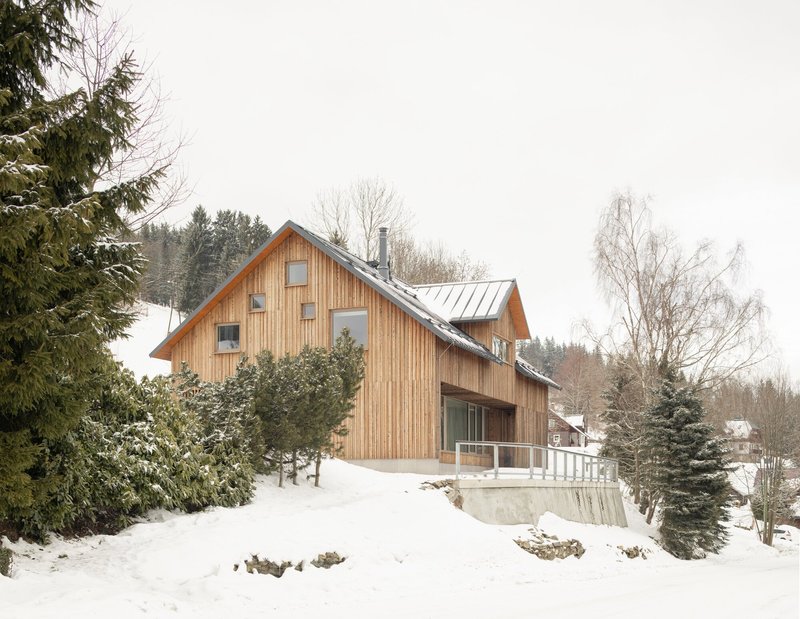 Front facade with vertical timber cladding and concrete terrace surrounded by snow-laden evergreens