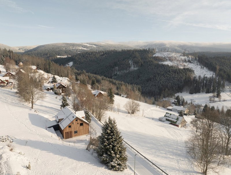 Aerial view of clustered timber houses on a snowy slope with forested hills beyond