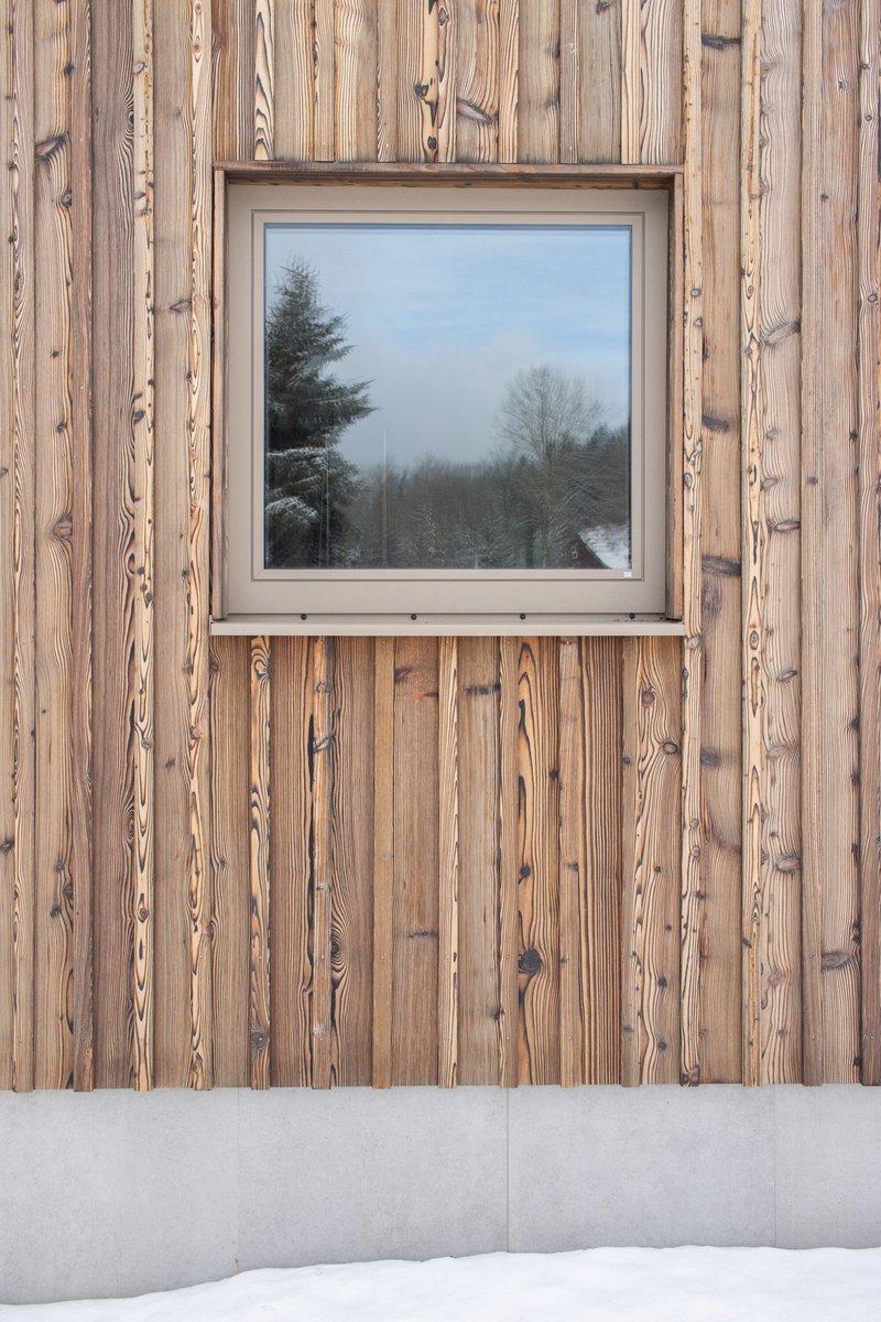 Square window set into weathered vertical timber cladding above a concrete base with snow drifts