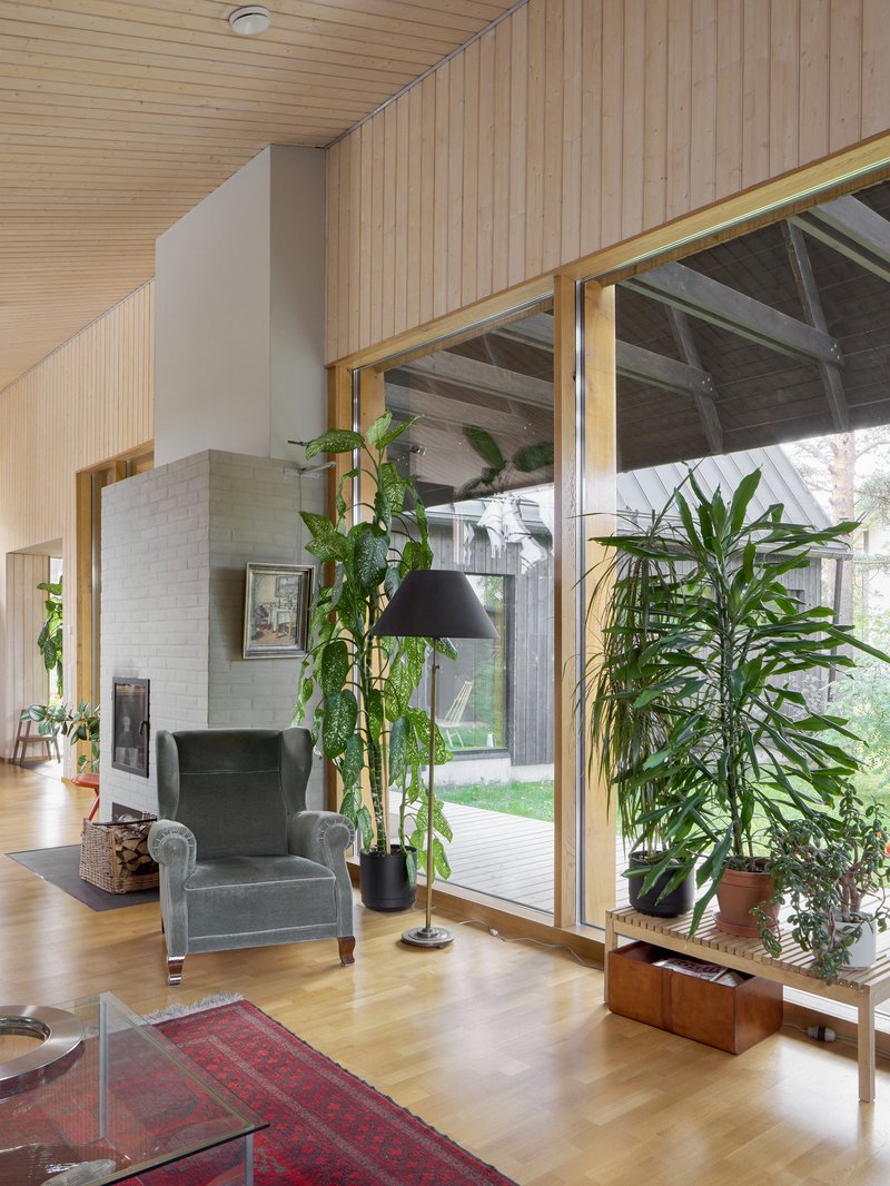 Living room with pale wood paneling, full-height glazing to courtyard and potted plants beside upholstered chair