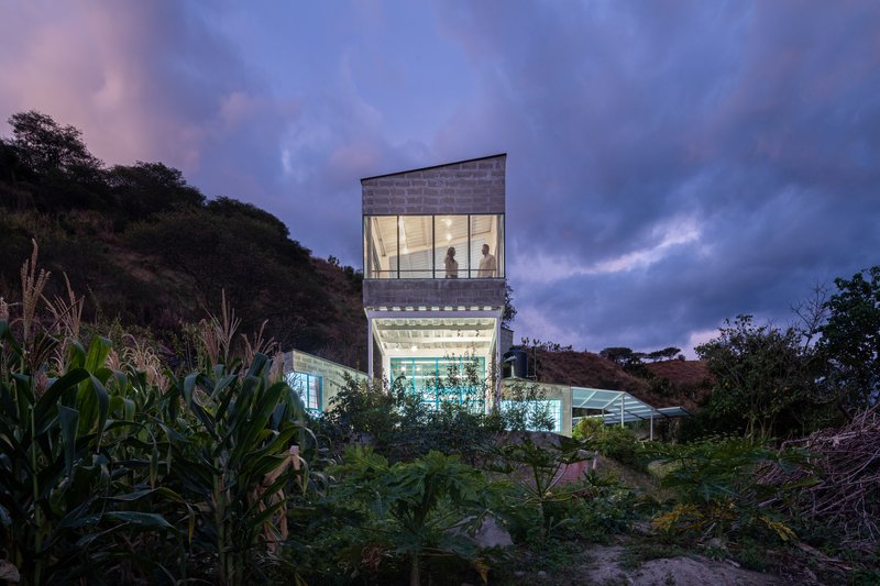 Illuminated tower volume at dusk with a figure visible through the transparent upper level