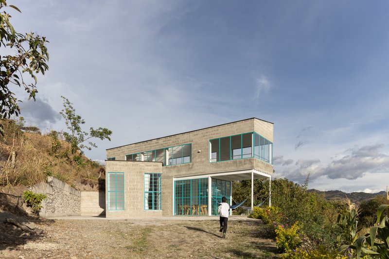 Two-story concrete block house with turquoise-framed windows and a covered terrace on a hillside clearing