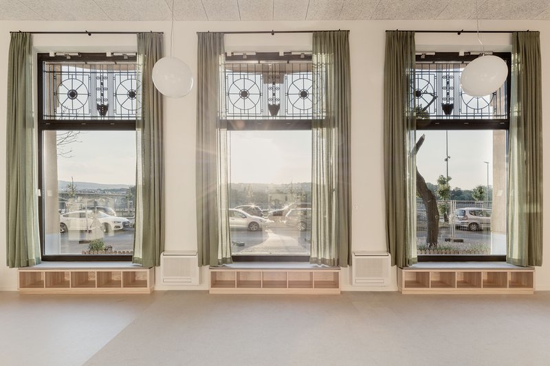 Three tall windows with geometric transom panels and sage curtains overlooking a parking lot in daylight