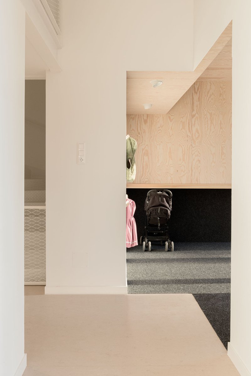 Plywood-clad alcove with integrated bench and dark carpet beside a white plaster corridor with a stroller
