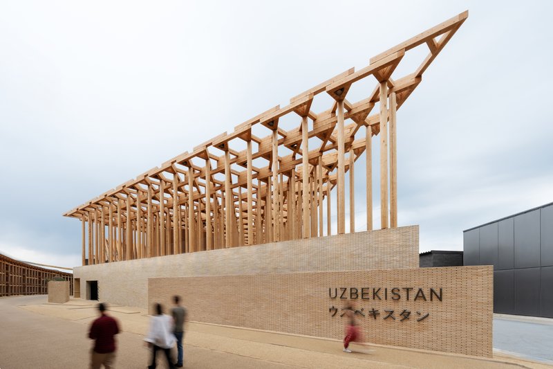 Diagonal timber roof structure rising above a beige brick facade with visitors walking past on an overcast day