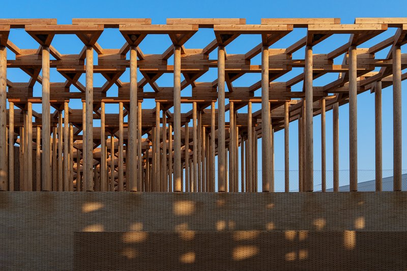 Timber beam ceiling structure above beige brick wall casting geometric shadows in bright blue sky