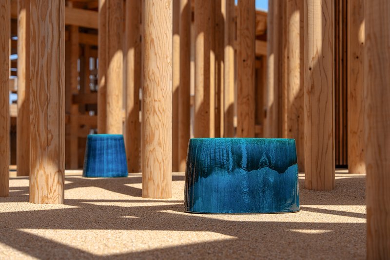 Blue glazed ceramic stools among timber columns casting shadows on gravel ground