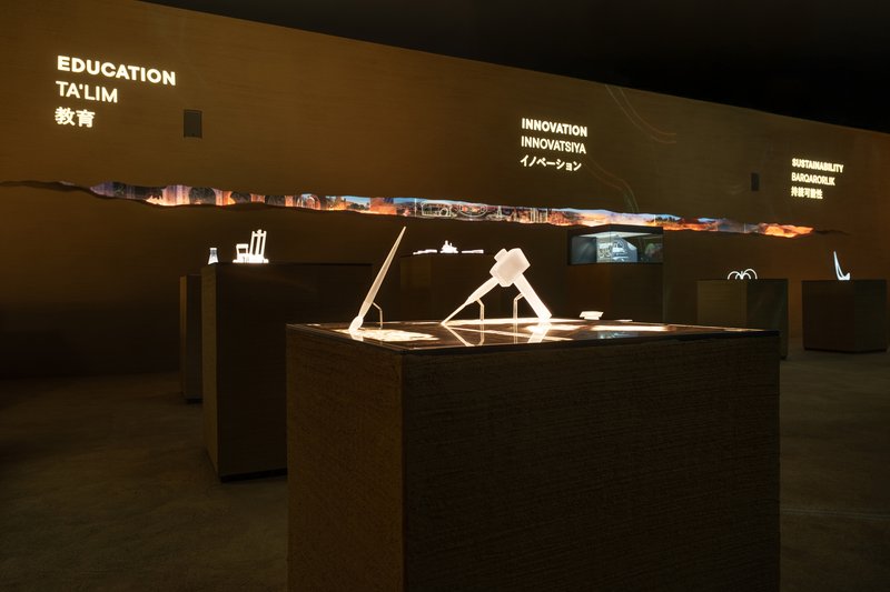 Exhibition tables with illuminated models beneath text panels in a darkened gallery space