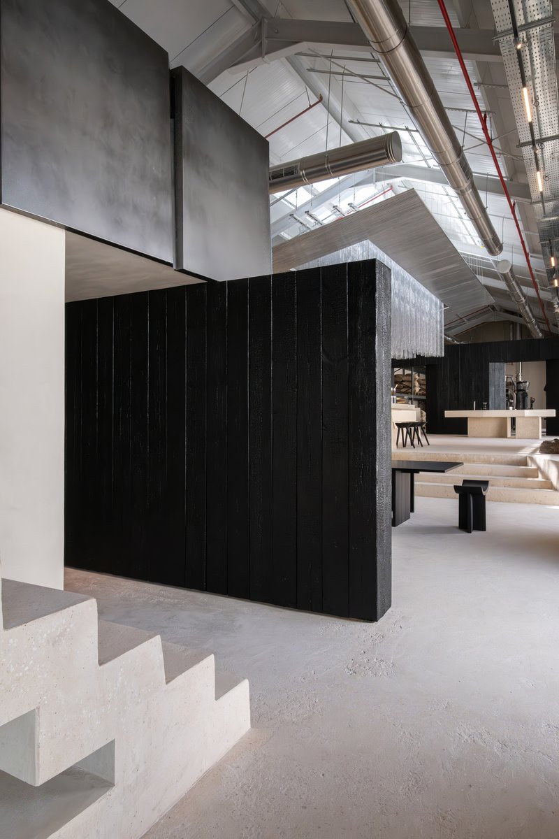 Charred timber partition beside pale stone staircase leading upward beneath exposed structural beams and red piping