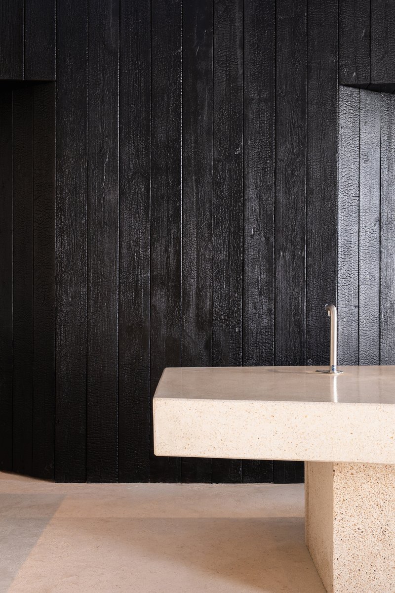 Terrazzo counter with cylindrical tap against charred timber cladding wall in darkened interior space