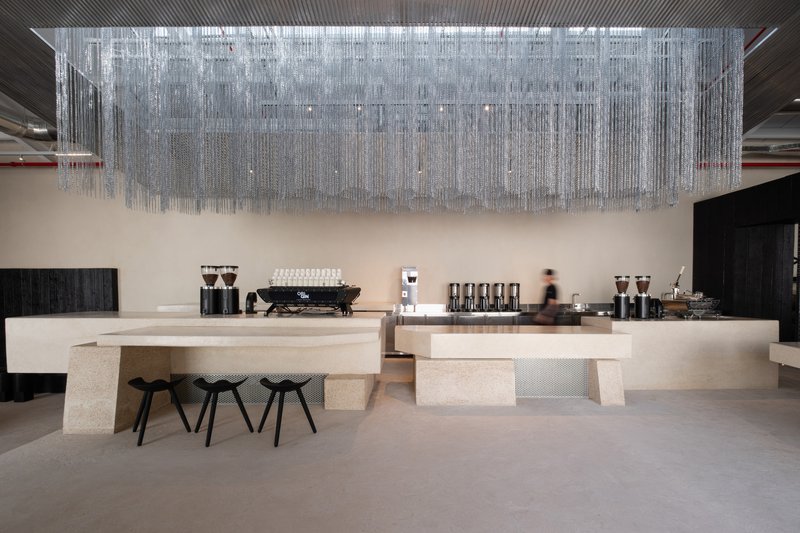 Long terrazzo service counter with espresso equipment beneath a cascading metal chain installation and a passing figure