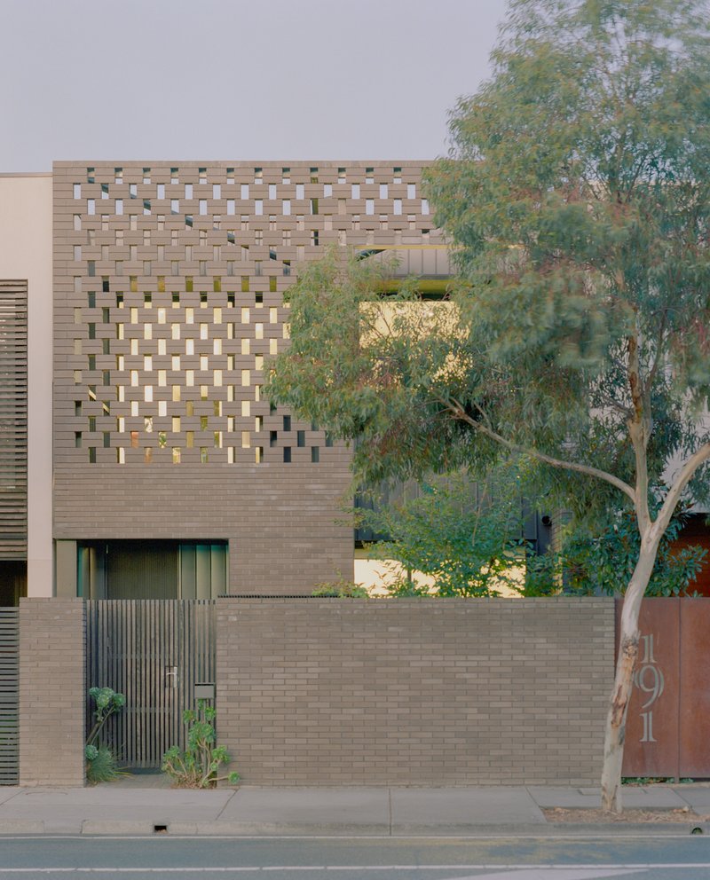 Perforated brick facade at dusk with street tree and internal lighting visible through openings