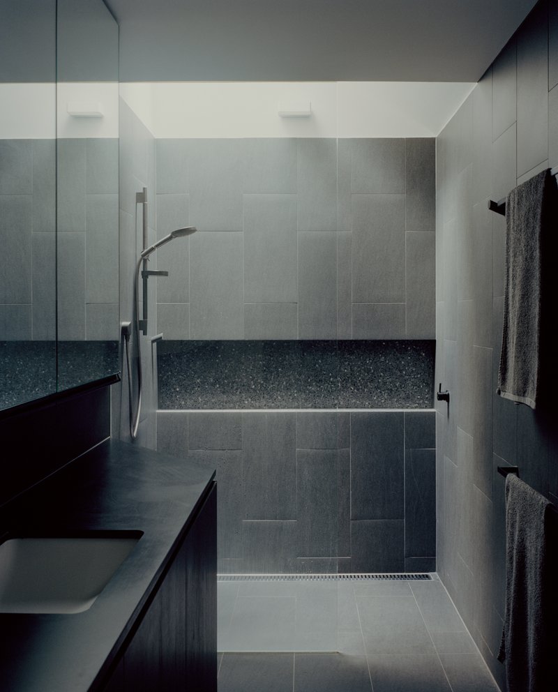 Walk-in shower enclosure with large-format grey tile walls and terrazzo bench under recessed ceiling lights