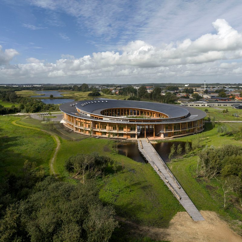 Aerial view of the ring-shaped structure with dark roof and timber colonnade approached by a wooden boardwalk