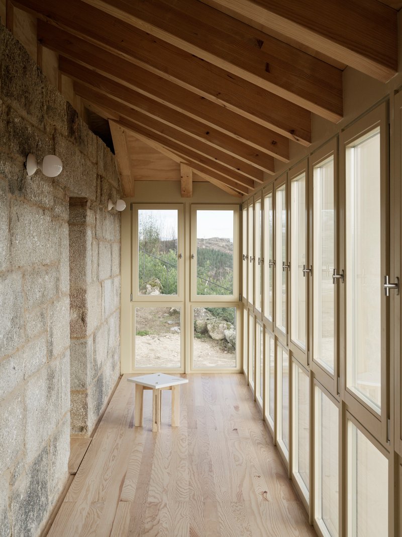Narrow glazed corridor with timber floor, exposed rafters and stone wall overlooking the landscape