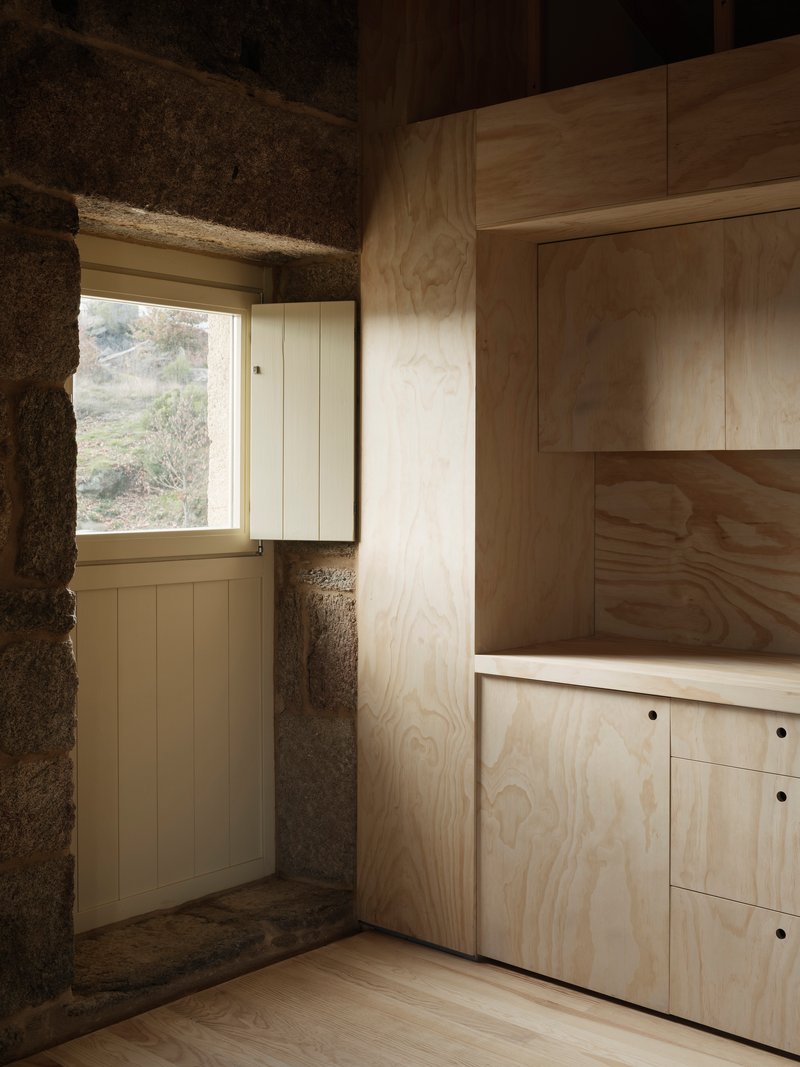 Corner window framed by exposed stone walls and plywood cabinetry with afternoon light streaming through