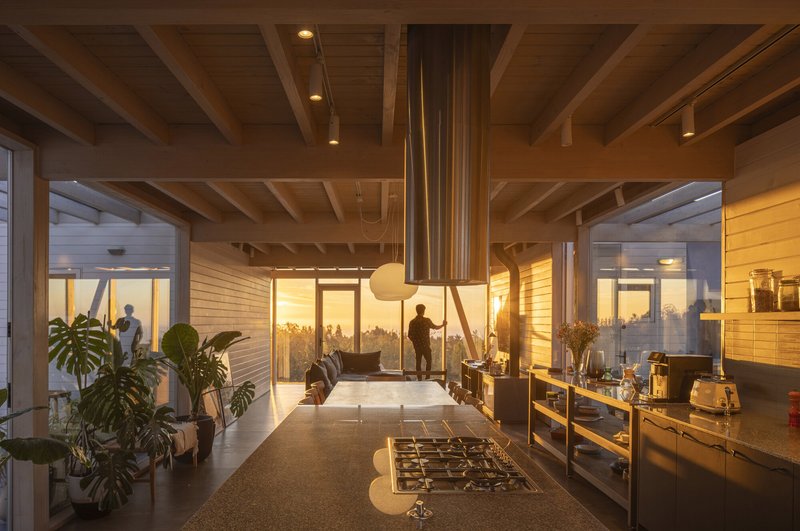 Kitchen with central island and cylindrical steel hood beneath exposed timber joists at dusk