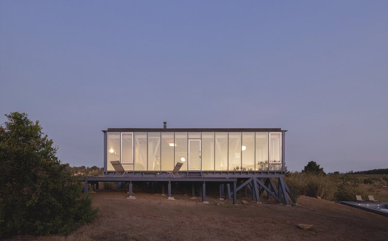 Glass-walled volume raised on steel trusses glowing with interior lighting at dusk