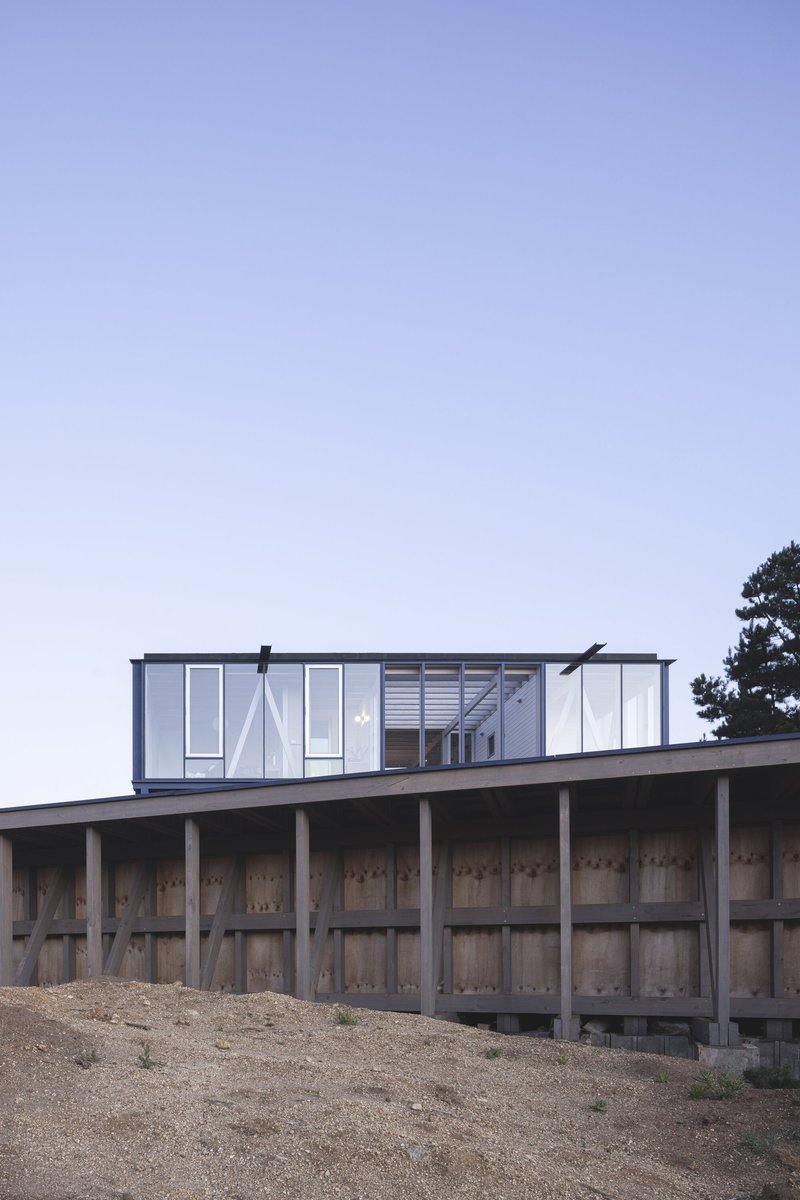 Elevated glass pavilion perched on exposed timber columns above a sloped gravel site