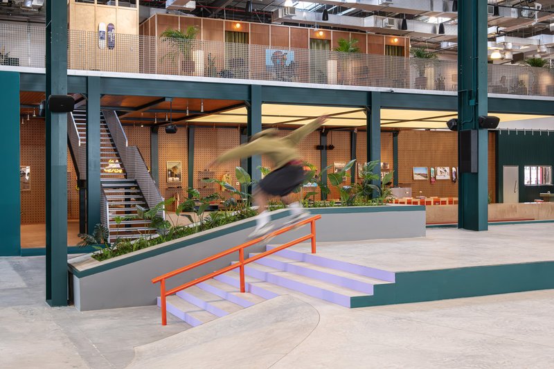 Double-height atrium with green steel columns, concrete skate ramp and a blurred skateboarder in motion